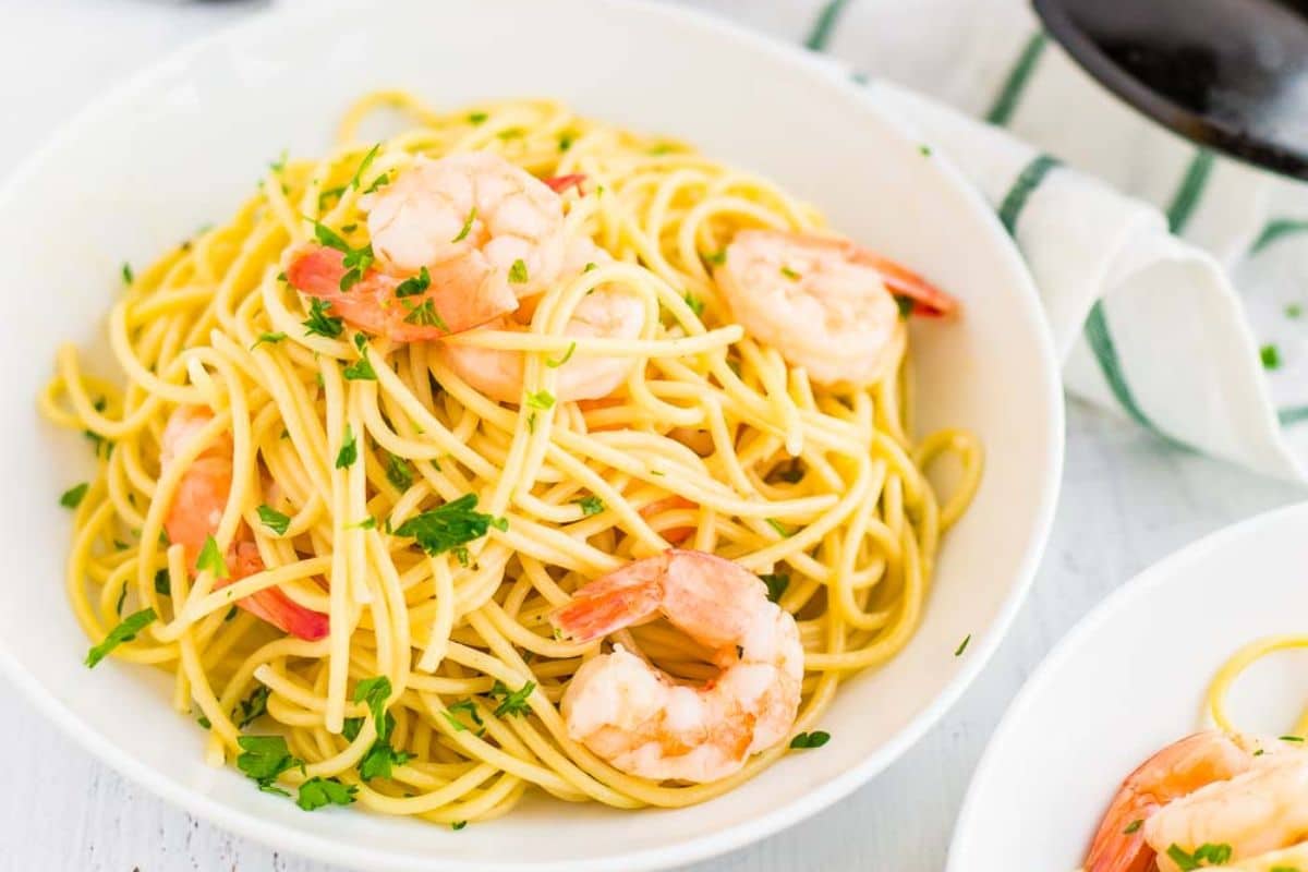 A bowl of spaghetti with shrimp, garnished with chopped parsley on a white table.