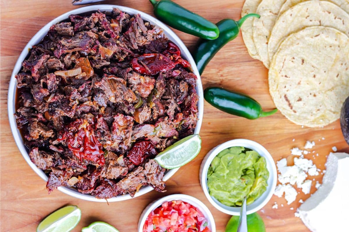 A bowl of shredded barbacoa with jalapeños, tortillas, guacamole, salsa, limes, and cheese on a wooden board.