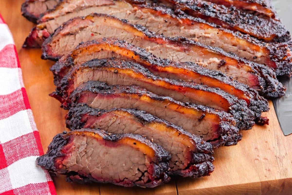 Sliced smoked brisket with a dark crust on a wooden board next to a red and white checkered cloth.