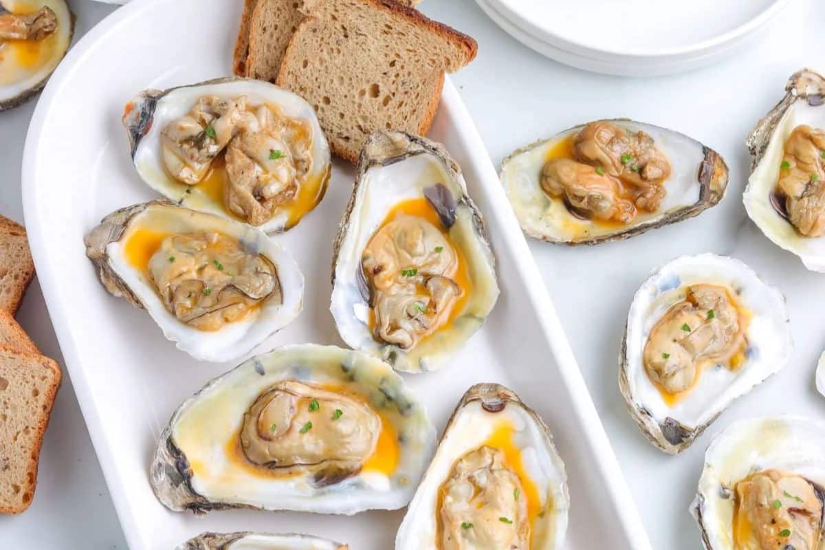 Oysters on the half shell topped with sauce, served with slices of brown bread on a white plate.