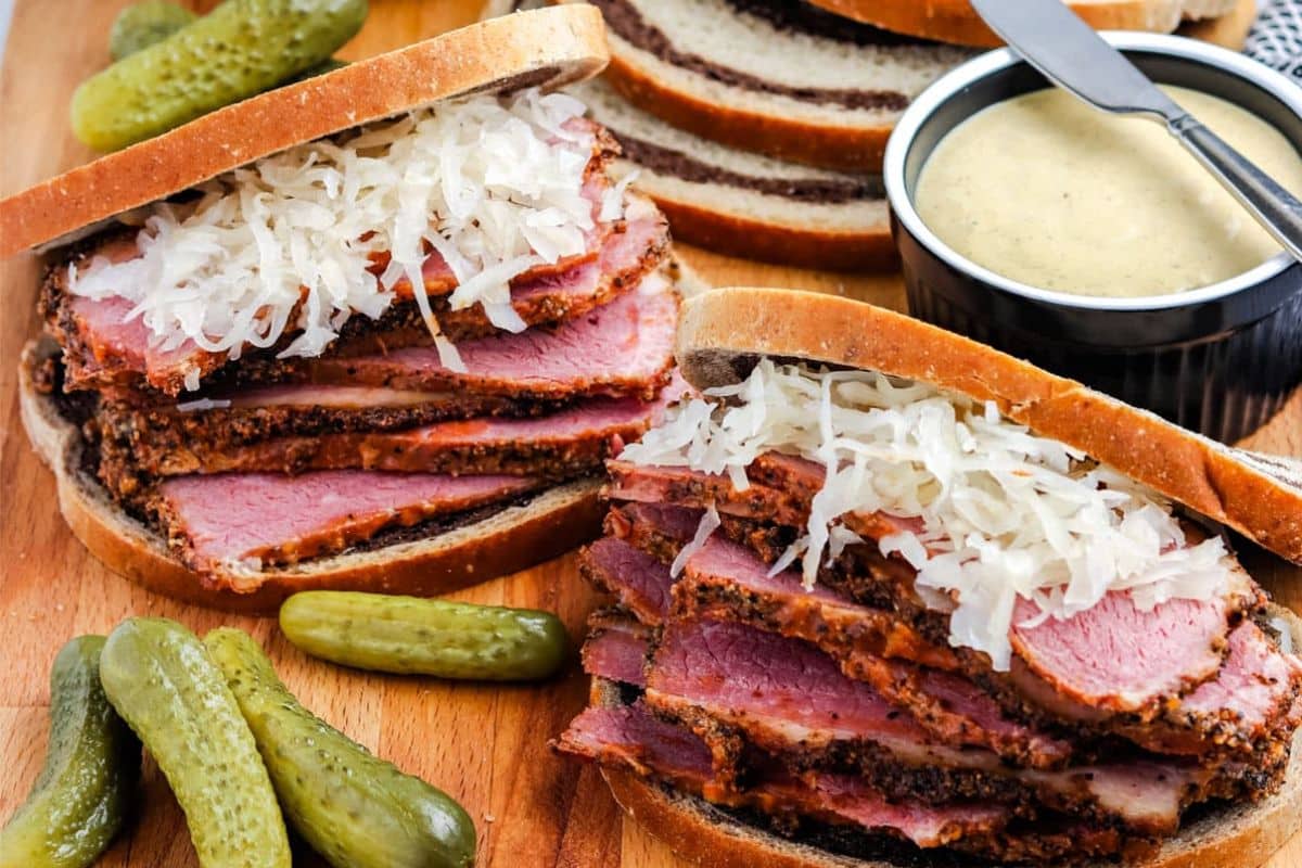 Two pastrami sandwiches with sauerkraut on rye bread, served with pickles and a bowl of mustard.