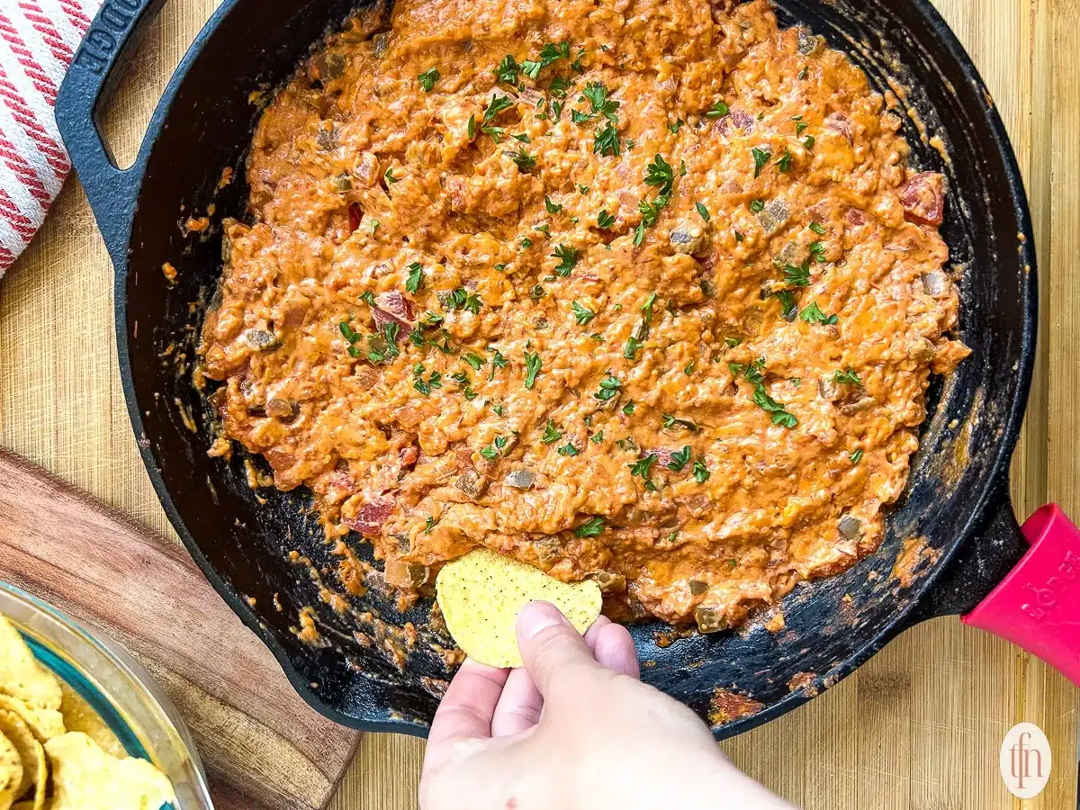 Hand dipping a tortilla chip into cheesy, tomato-based dip in a black skillet on a wooden surface.