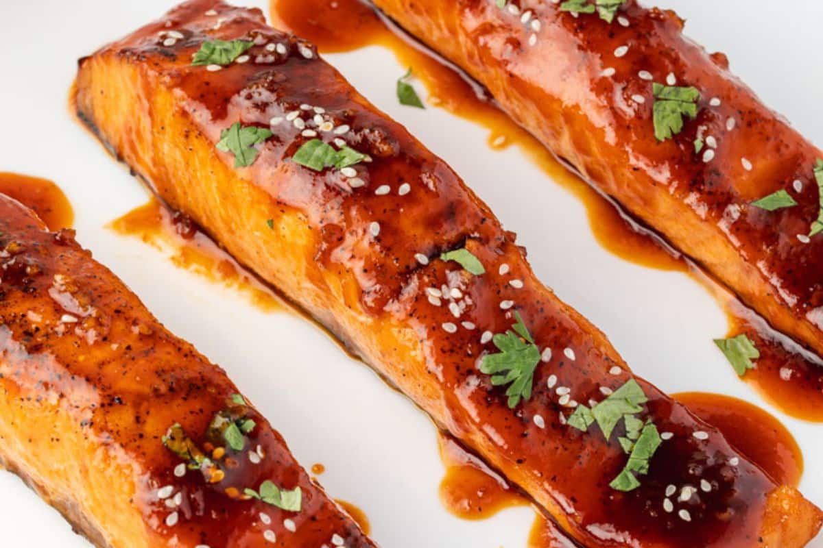 Three glazed salmon fillets topped with sesame seeds and chopped herbs on a white plate.