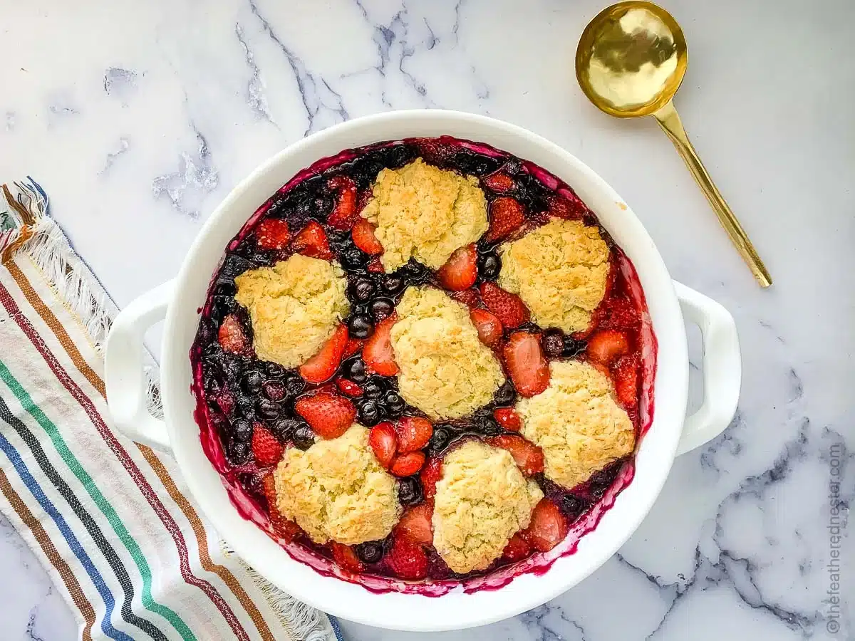 A berry cobbler with golden biscuit topping in a white dish, next to a gold spoon on a marble surface.