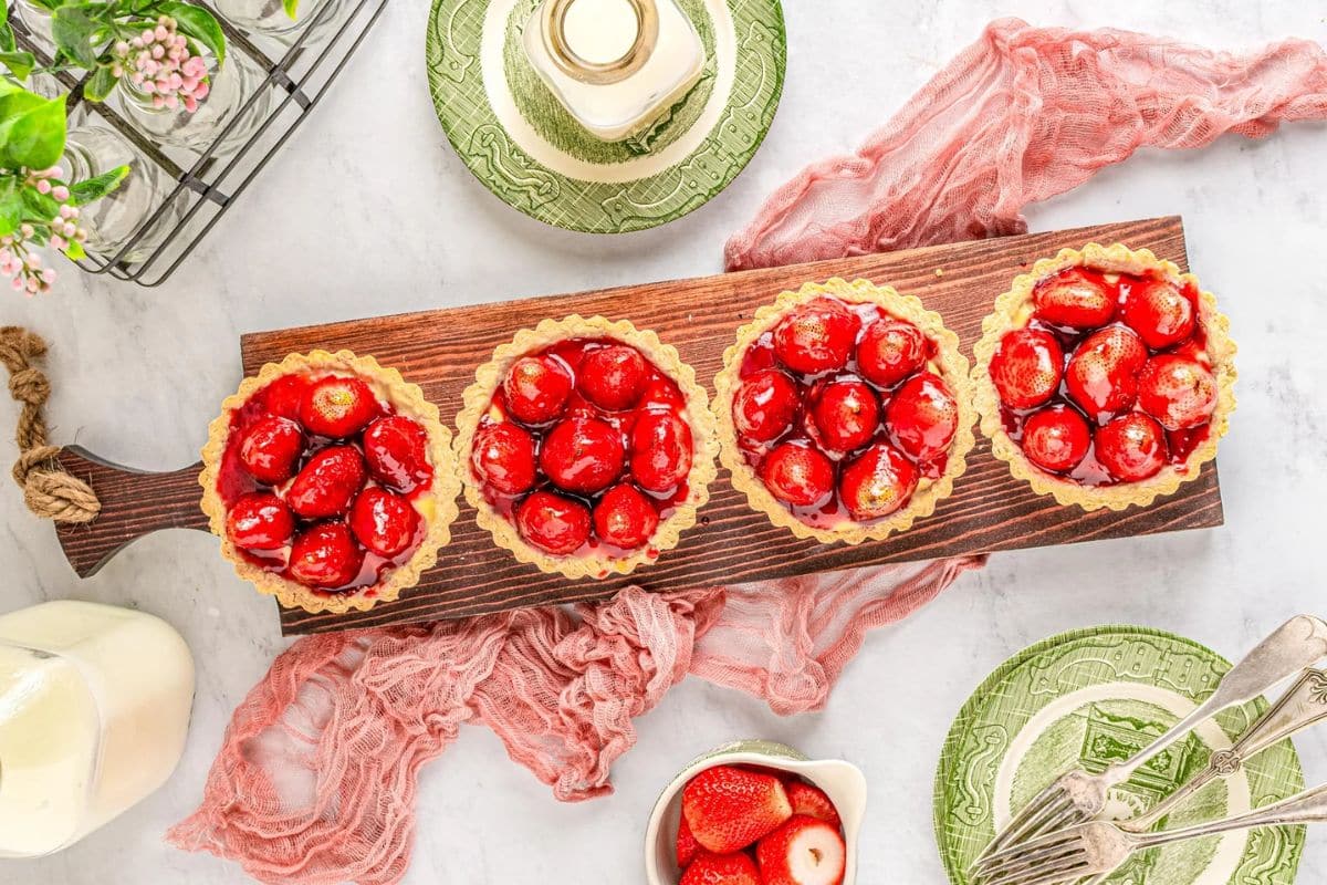 Four strawberry tarts on a wooden board, surrounded by plates, strawberries, milk, and cutlery on a table.