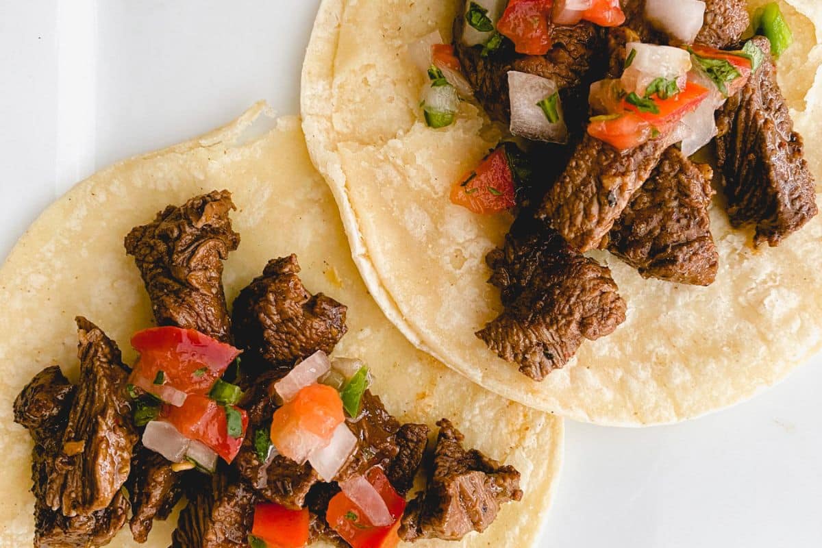 Two corn tortillas topped with grilled steak pieces and fresh tomato salsa on a white plate.