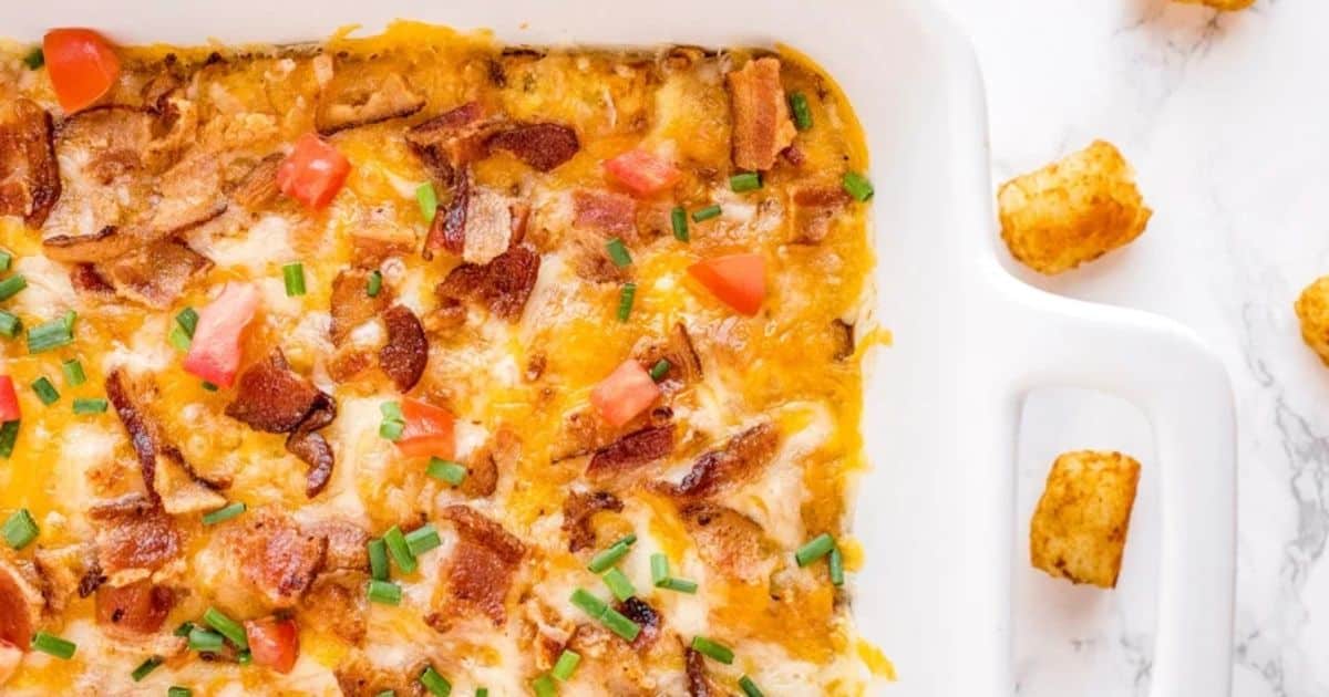 Casserole topped with melted cheese, bacon, chopped tomatoes, and chives in a white baking dish with tater tots nearby.