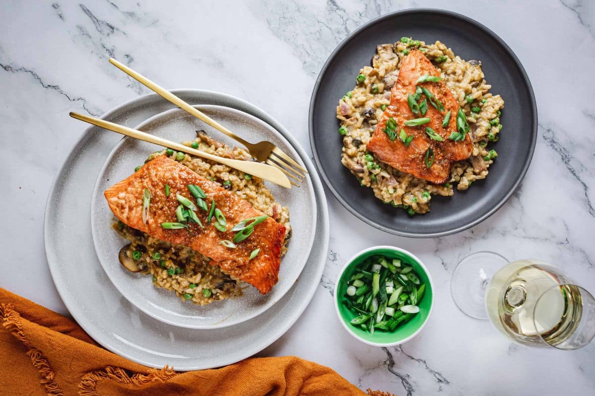 Two plates of salmon over risotto, topped with green onions, with a bowl of onions and a glass of white wine.