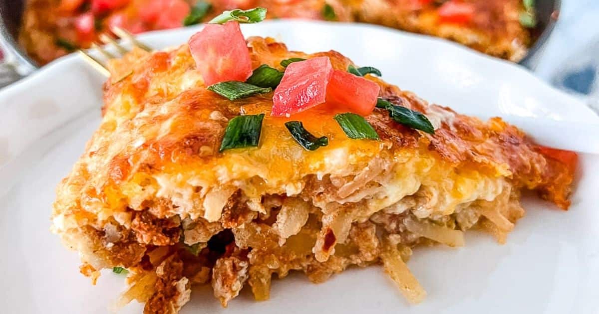 A slice of cheesy casserole topped with chopped tomatoes and green onions on a white plate.