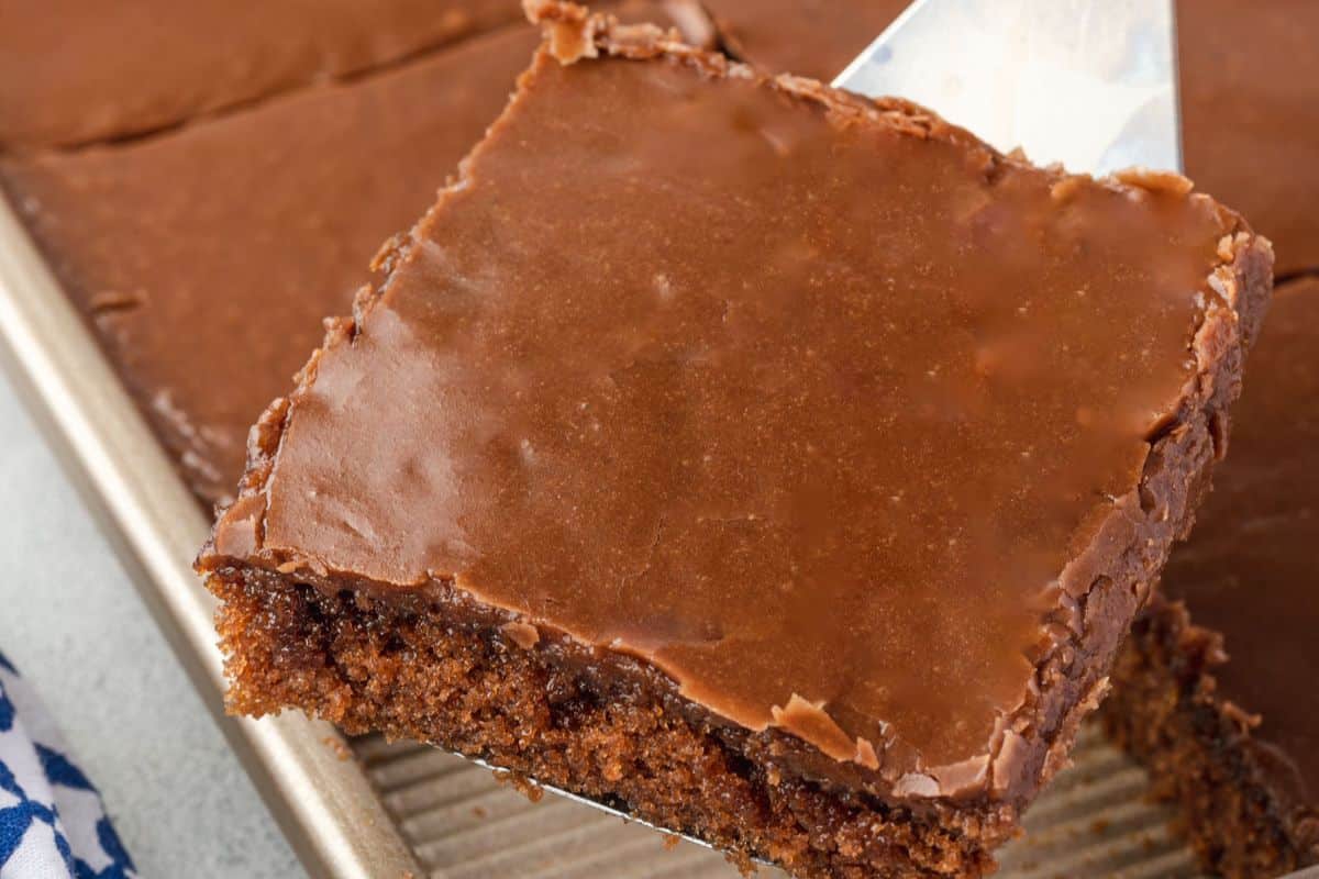 A close-up of a chocolate sheet cake square being lifted from a pan with a spatula.