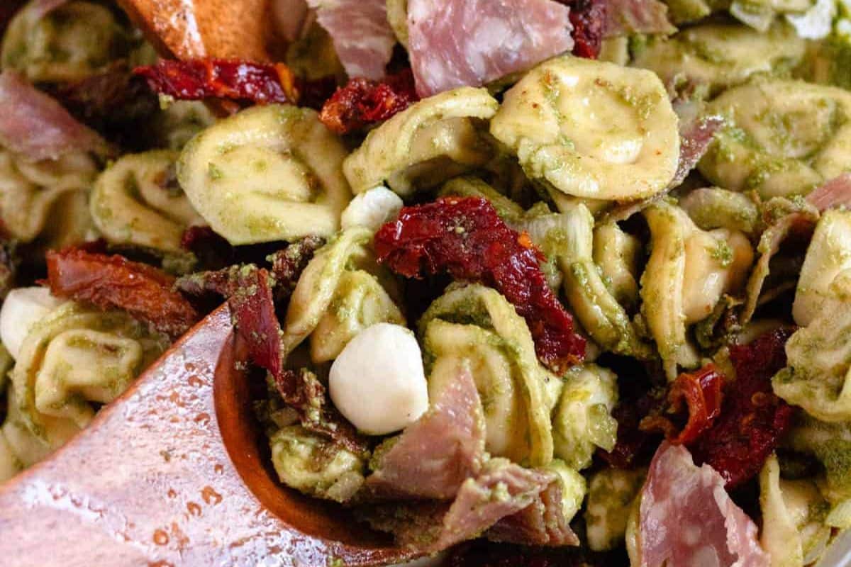 Orecchiette pasta with pesto, sun-dried tomatoes, mozzarella, and prosciutto, mixed with wooden utensils.