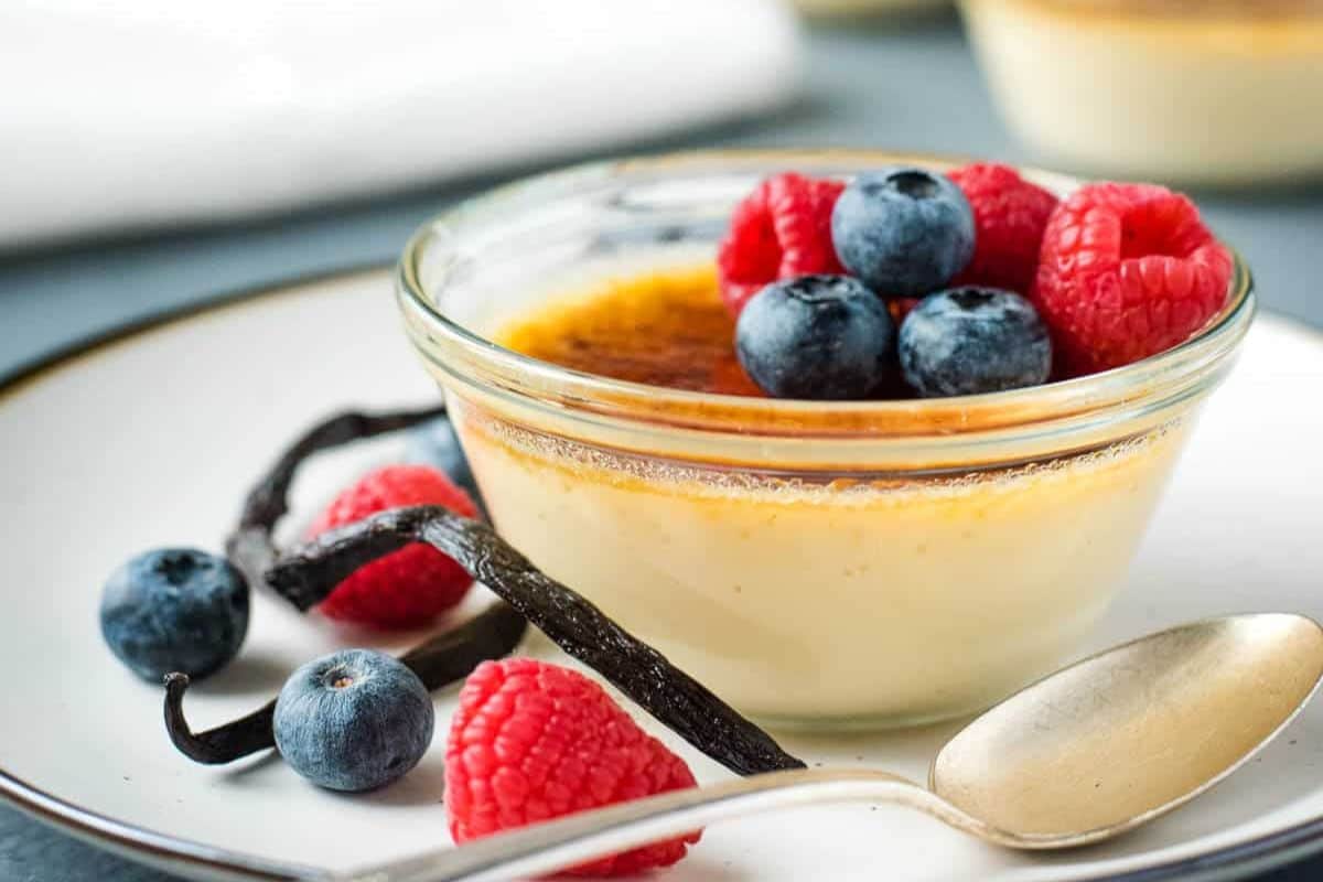 Glass bowl of crème brûlée topped with raspberries and blueberries, with vanilla beans on the side.