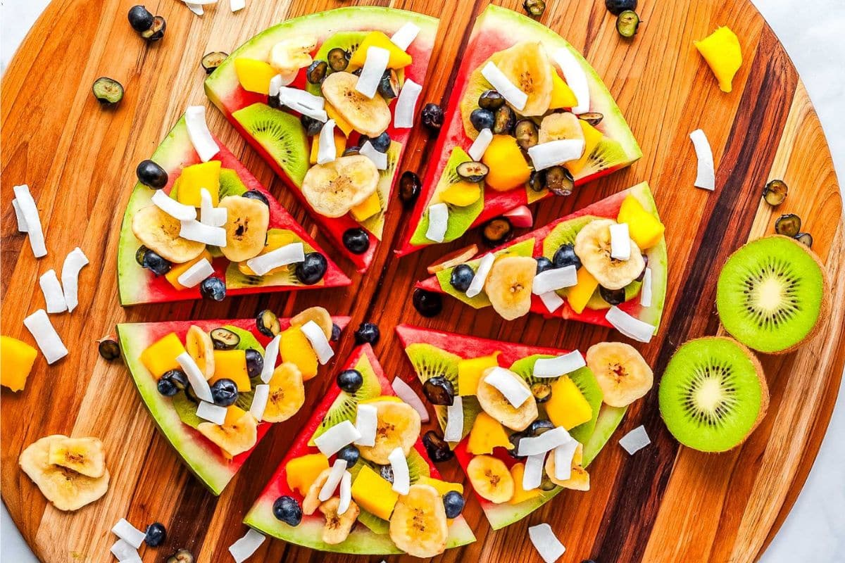 Sliced watermelon topped with banana, mango, blueberries, coconut flakes, and kiwi on a wooden board.