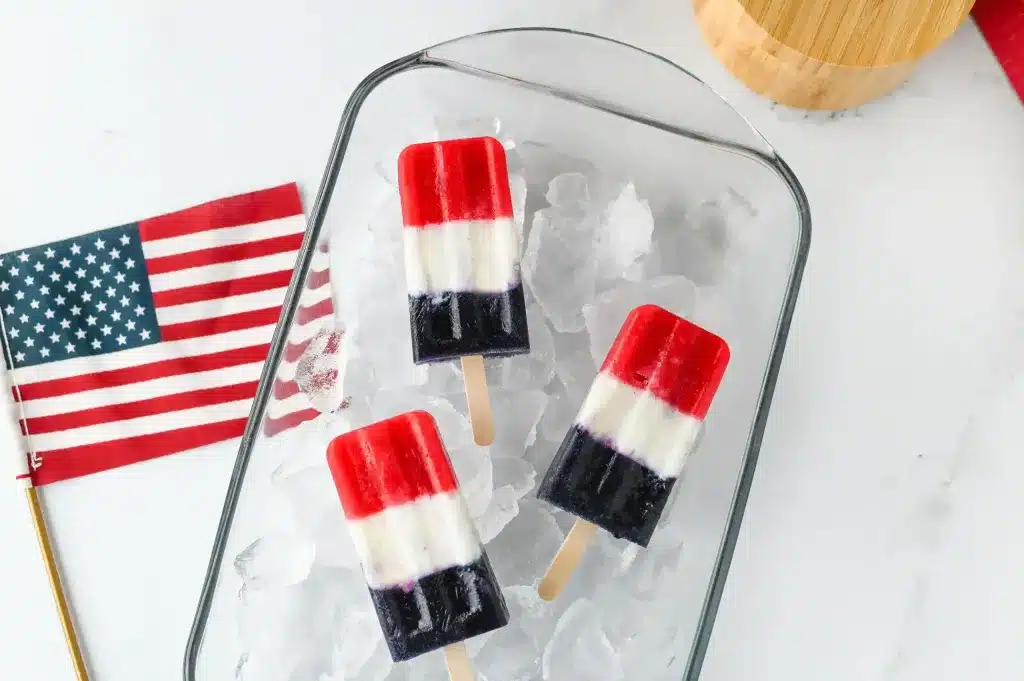 Three red, white, and blue popsicles on ice in a glass dish next to a small American flag.