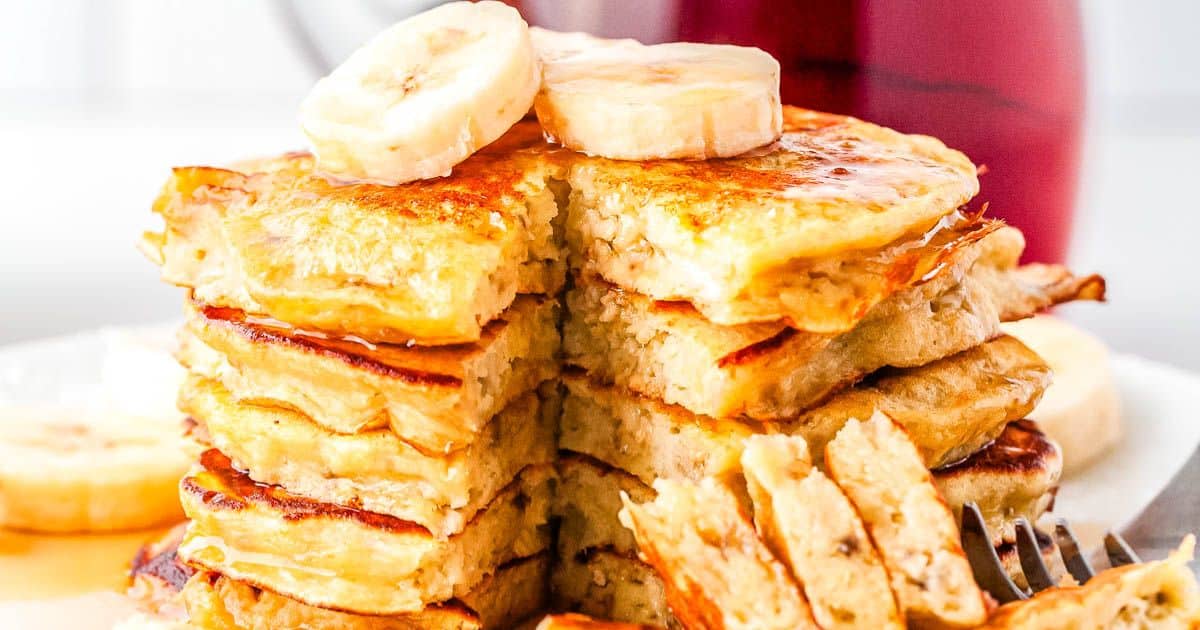 A stack of banana pancakes topped with banana slices and syrup, with a bite cut out on a white plate.