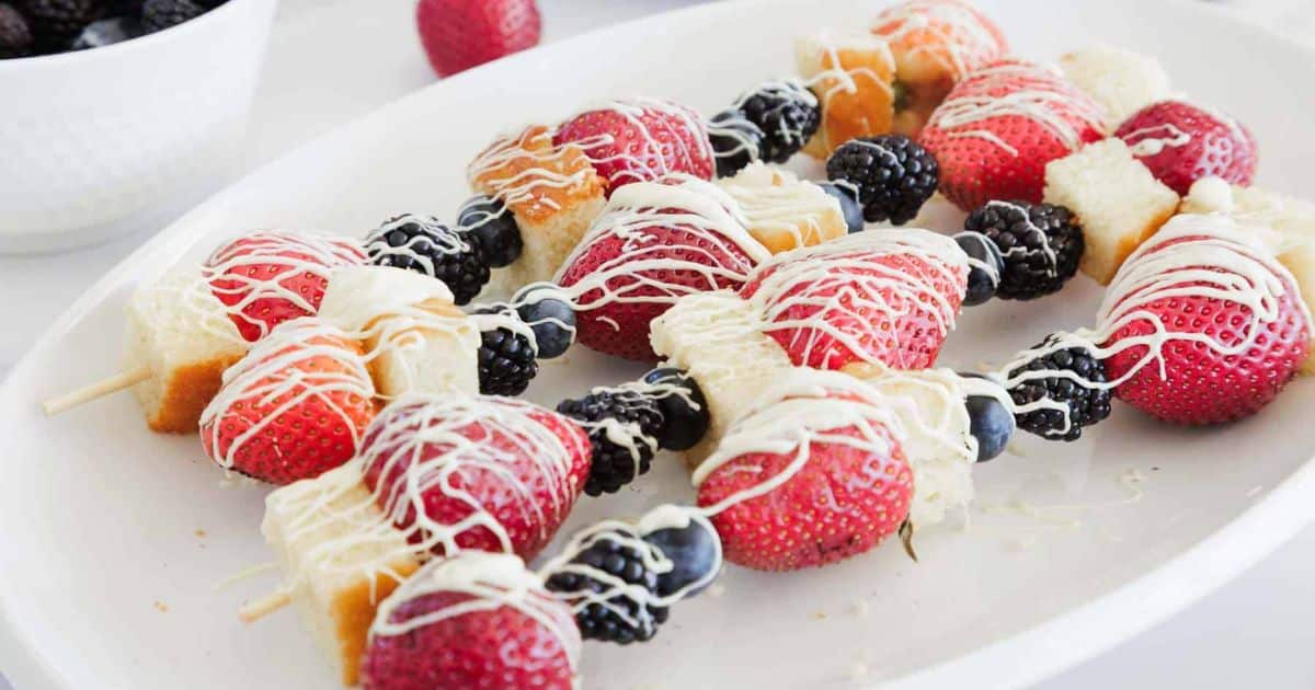 Fruit and cake skewers drizzled with white chocolate, featuring strawberries, blackberries, and blueberries.