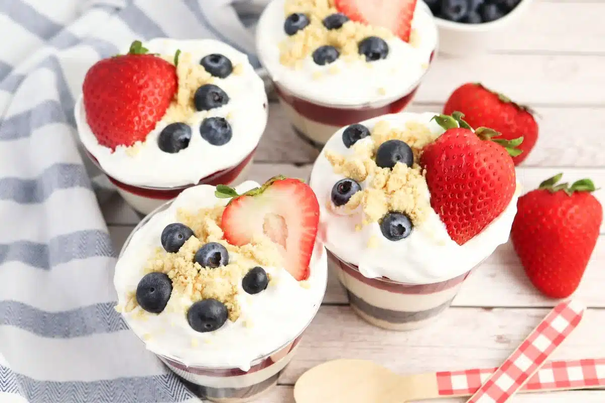 Four dessert cups with whipped cream, blueberries, and strawberries, topped with crumbly topping on a wooden table.