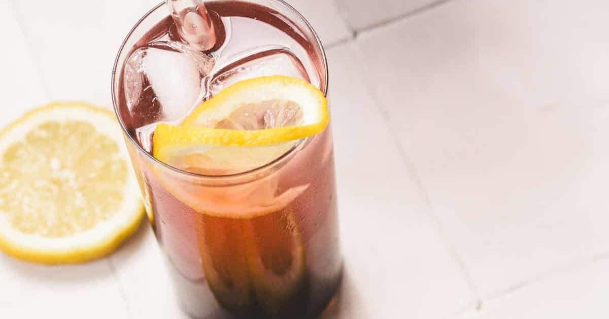 A glass of iced tea with a lemon slice and a straw, next to a slice of lemon on a white surface.