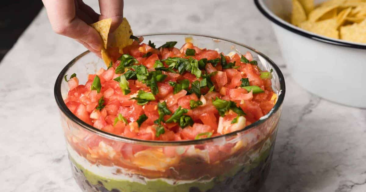 A hand dips a tortilla chip into a layered dip topped with diced tomatoes and herbs.
