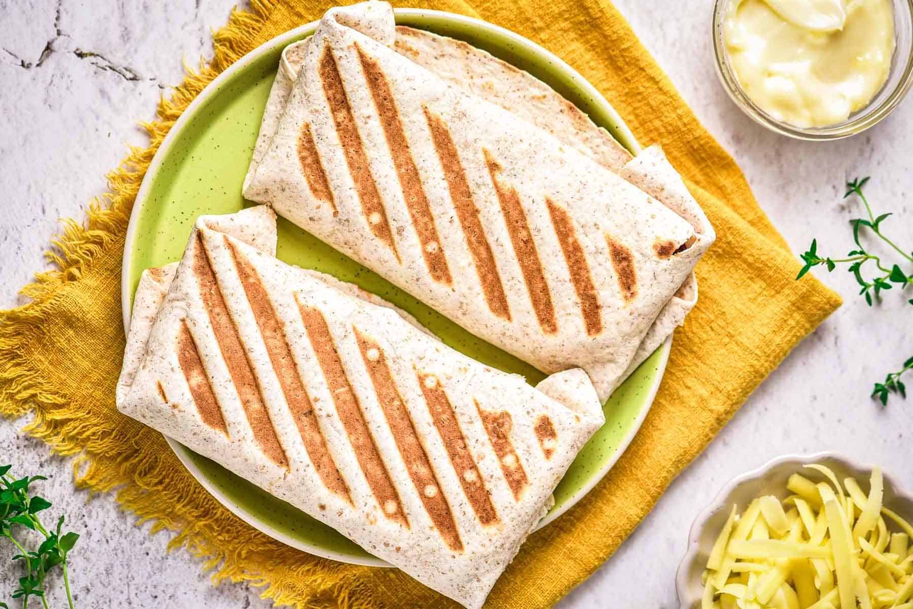 Two grilled wraps with golden grill marks on a green plate, set on a yellow cloth with cheese and dip nearby.