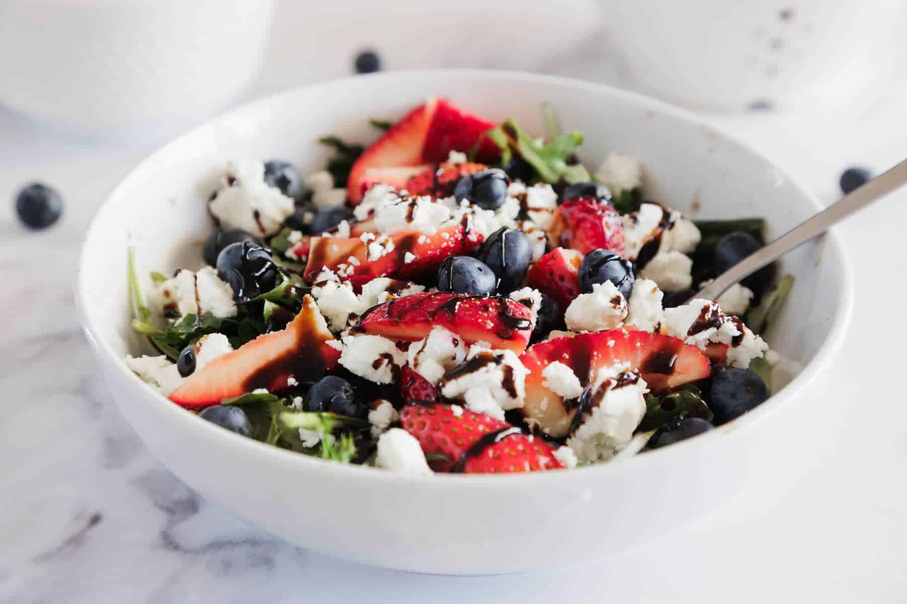 A bowl of salad with strawberries, blueberries, feta cheese, and balsamic glaze on a white surface.