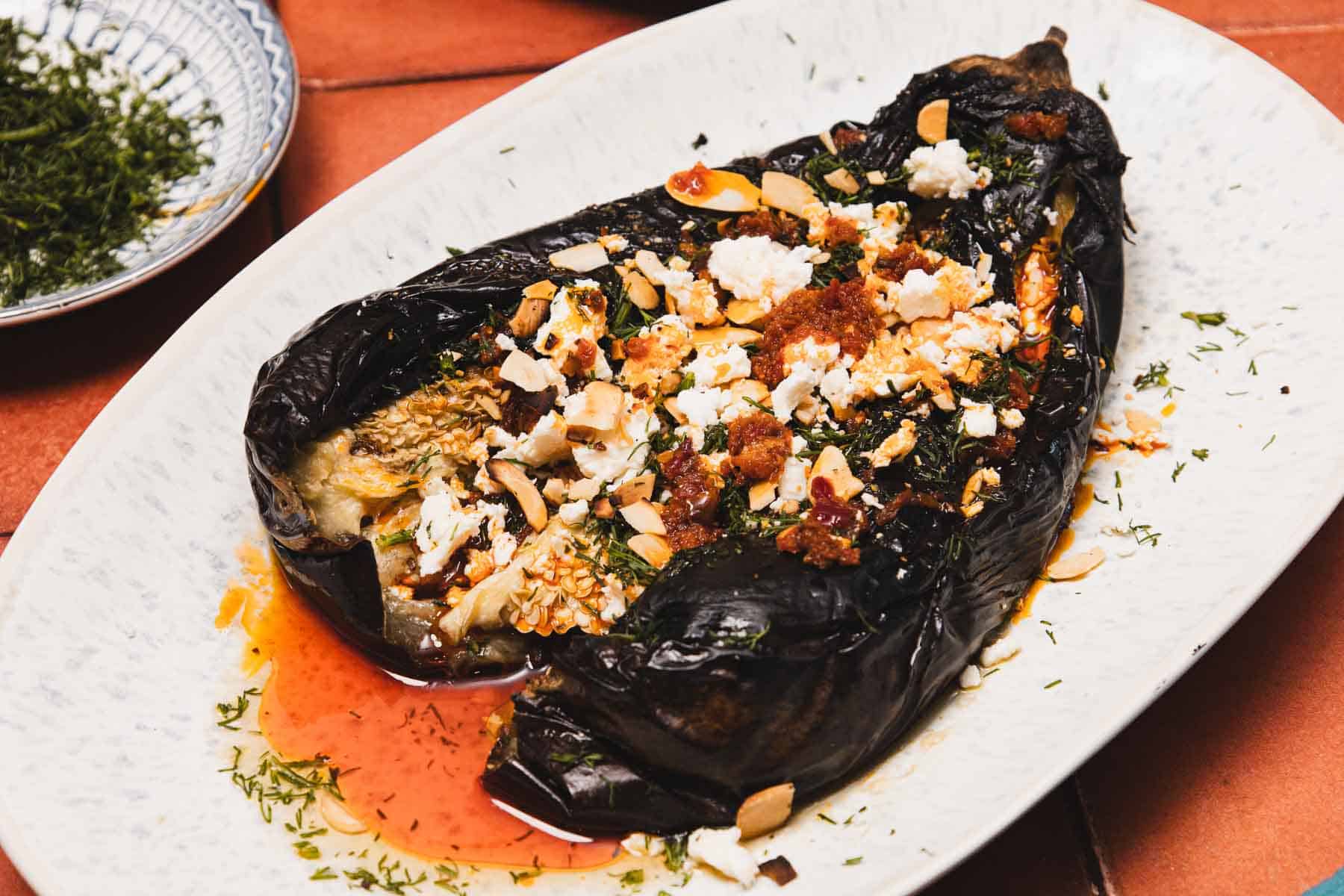 Roasted eggplant topped with crumbled cheese, nuts, herbs, and sauce on a white oval plate.