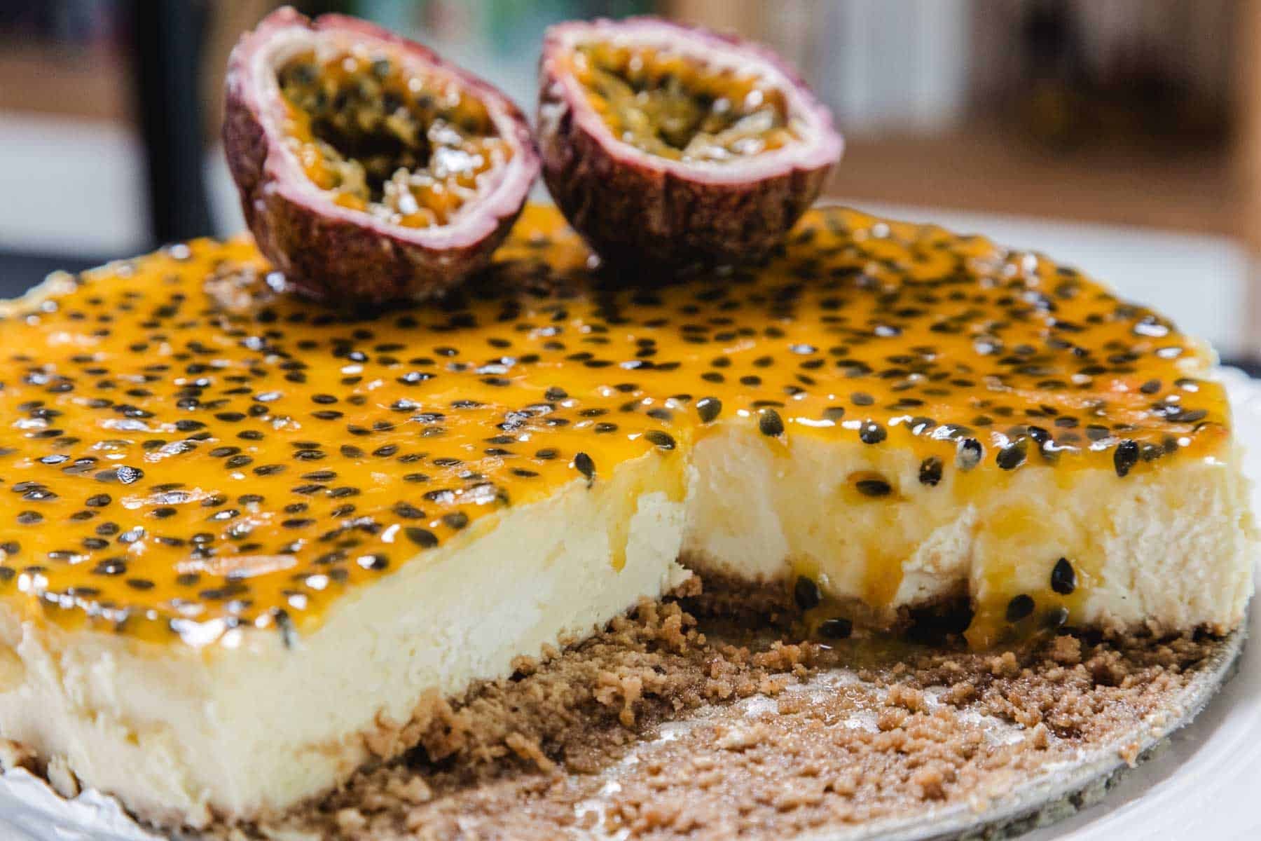 A cheesecake topped with passion fruit and seeds, with a slice removed, showing a crumbly base.