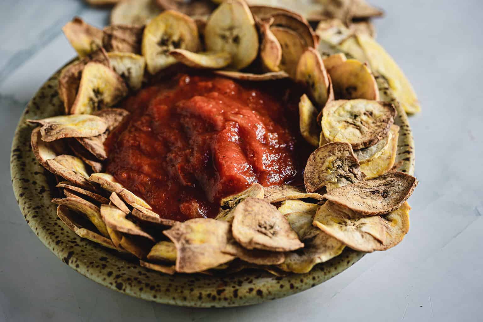 A bowl of plantain chips surrounds a bowl of red tomato salsa on a speckled plate.