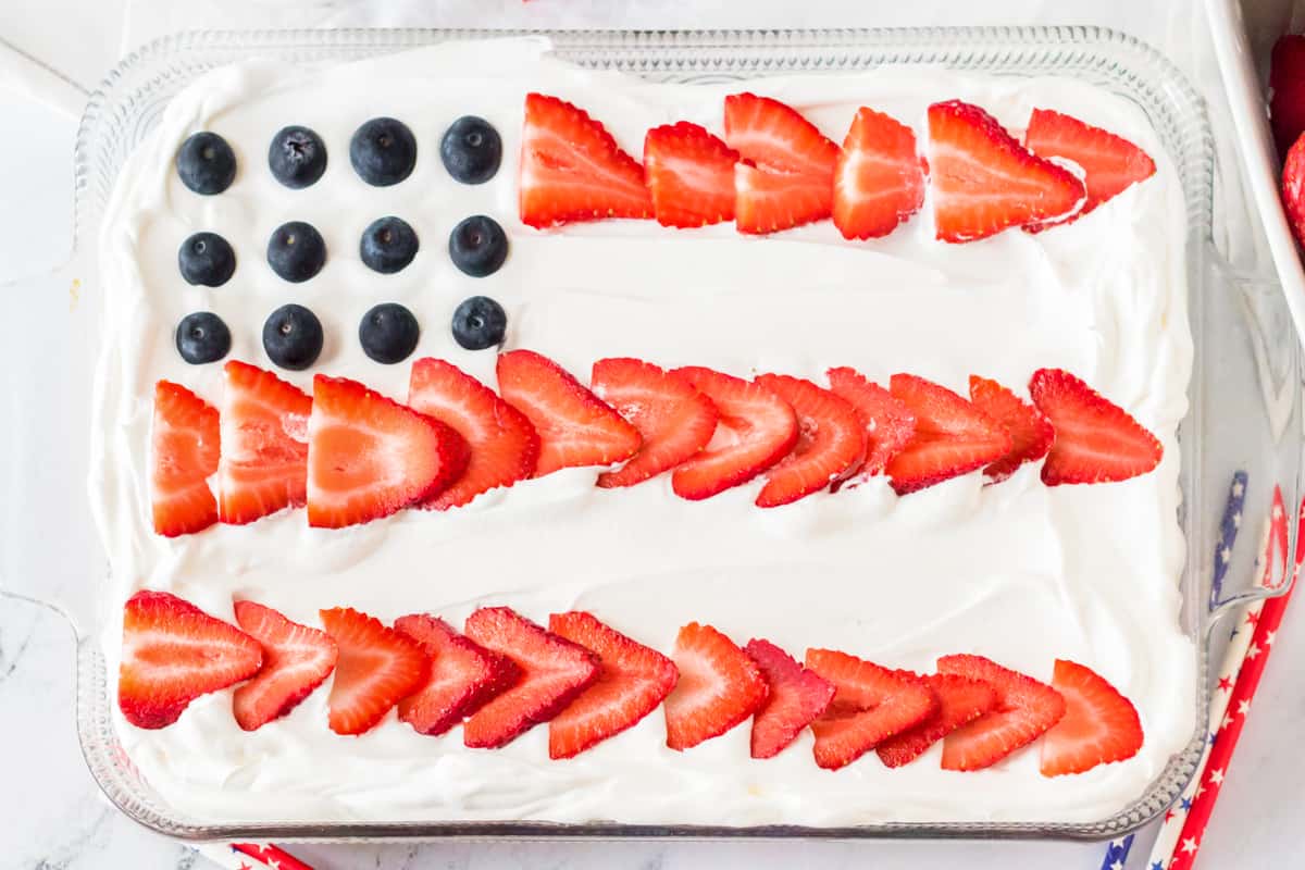 Rectangular cake with white frosting, topped with strawberry stripes and blueberries arranged like an American flag.