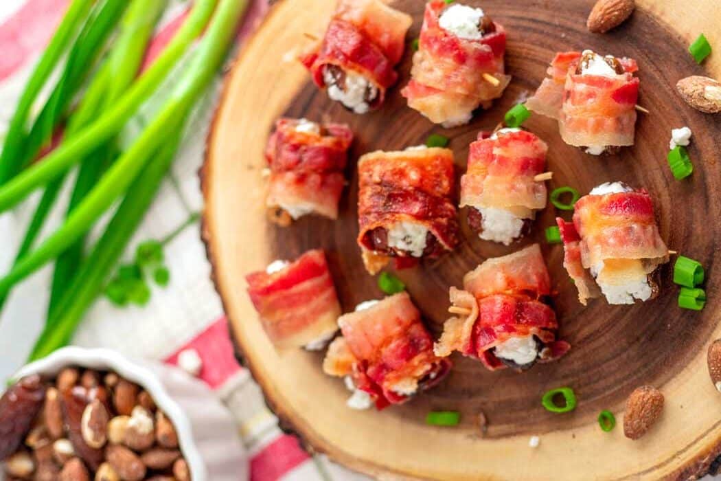 Bacon-wrapped stuffed dates with goat cheese on a wooden serving board, garnished with green onions.