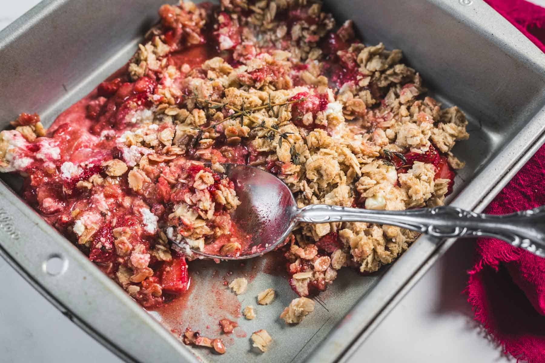 A partially served strawberry crumble in a metal baking dish with a spoon resting inside.