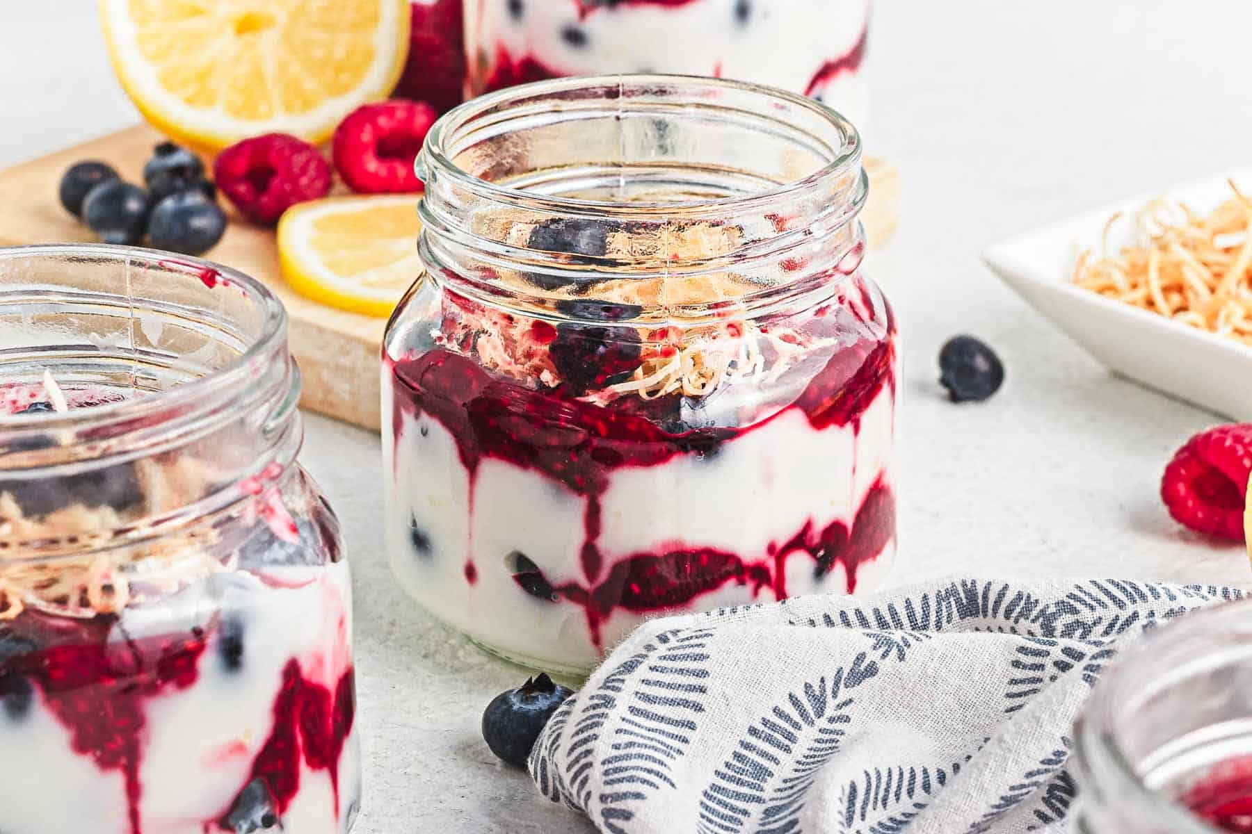Glass jars filled with yogurt, berry compote, toasted coconut, and fresh fruit on a light surface.
