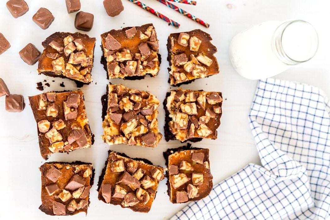 Nine chocolate and caramel dessert bars on a white surface with a checkered towel and a bottle of milk.