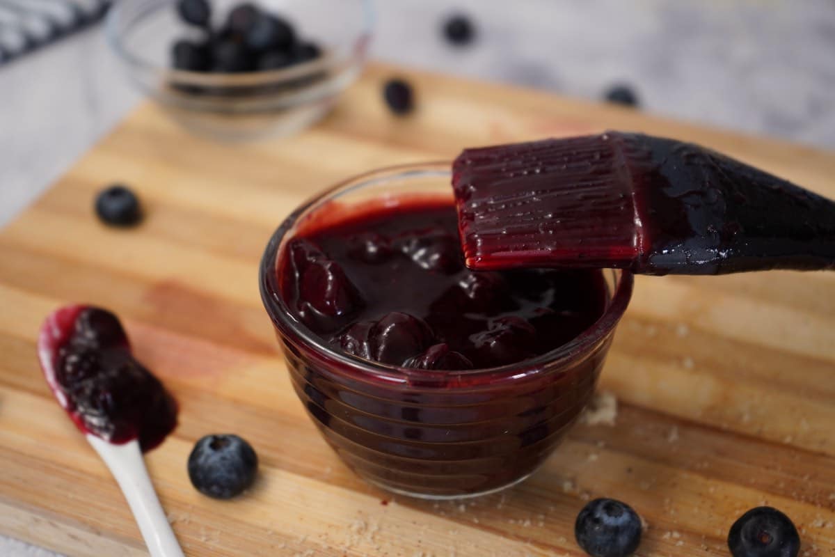 A glass bowl of blueberry sauce with a basting brush on a wooden board, fresh blueberries nearby.
