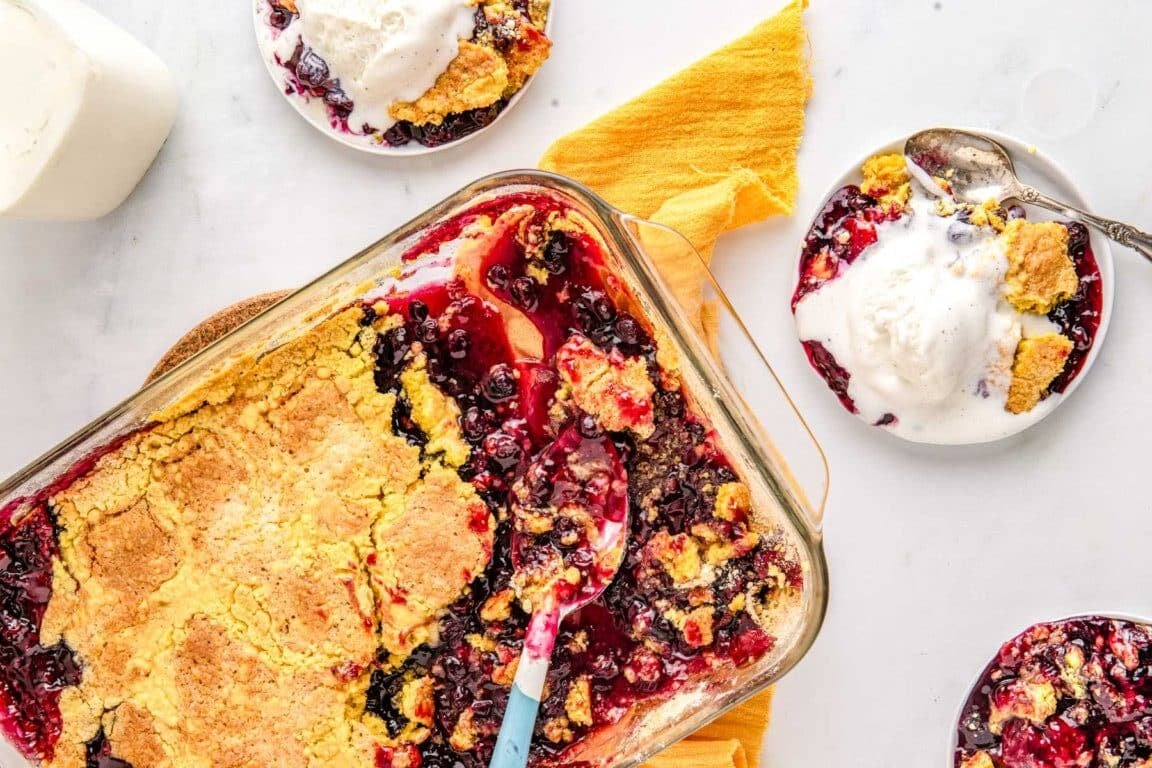 A baking dish of berry cobbler with a golden crust, served with vanilla ice cream on small plates nearby.
