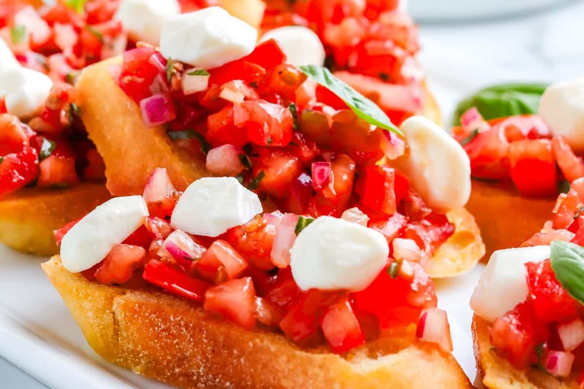 Toasted bread topped with chopped tomatoes, mozzarella, red onion, and basil on a white plate.