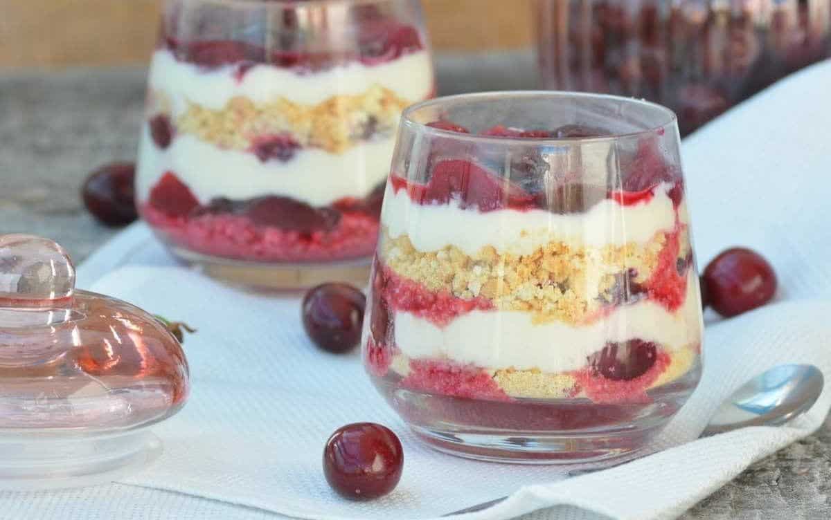 Dessert glasses with layers of cream, cherries, and crumbs on a white napkin, surrounded by fresh cherries.