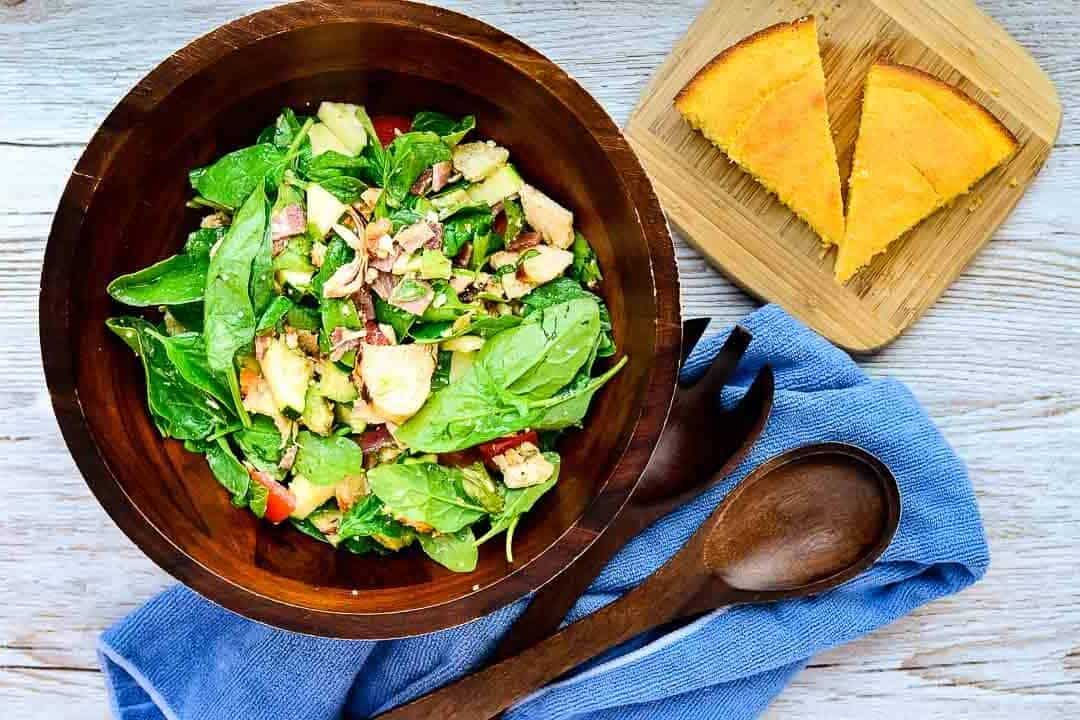 A wooden bowl of spinach salad with apples and nuts, next to two cornbread slices on a wooden plate.