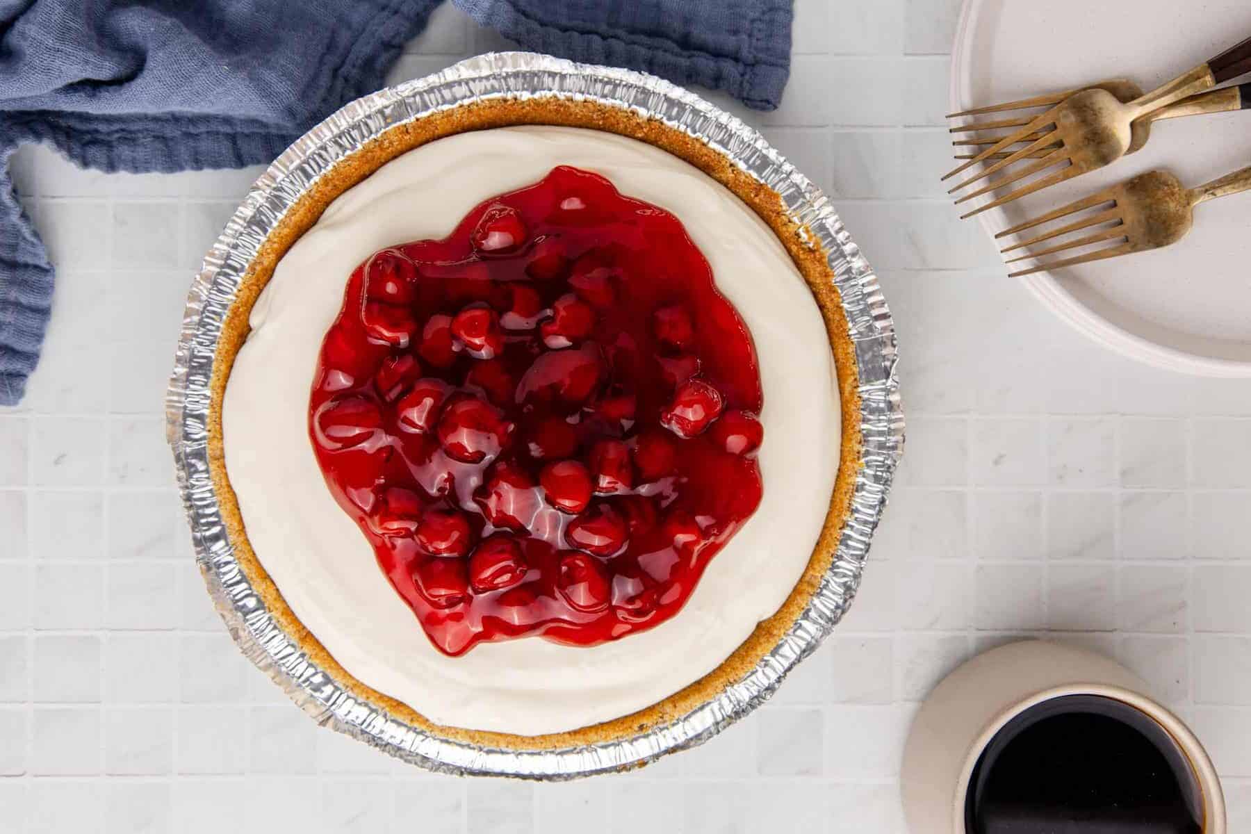 Cherry cheesecake with a graham crust in a foil pan, topped with cherry filling and whipped cream.
