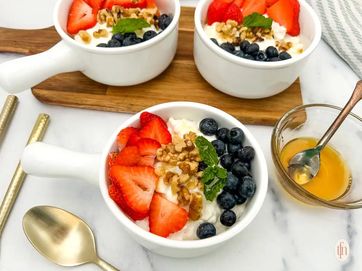 Three bowls of yogurt topped with strawberries, blueberries, walnuts, and mint, with honey and spoons nearby.