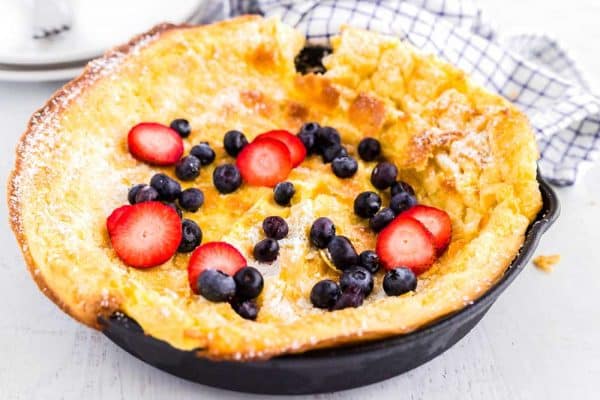 A Dutch baby pancake in a skillet topped with sliced strawberries, blueberries, and powdered sugar.
