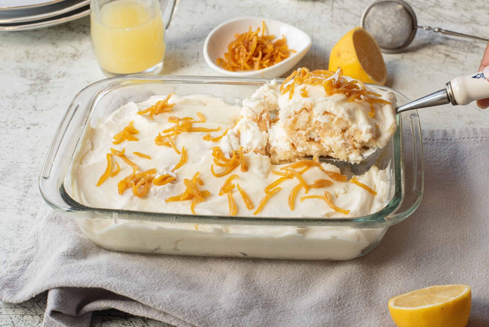 Glass dish with creamy lemon dessert topped with zest; a serving is being scooped out with a spatula.