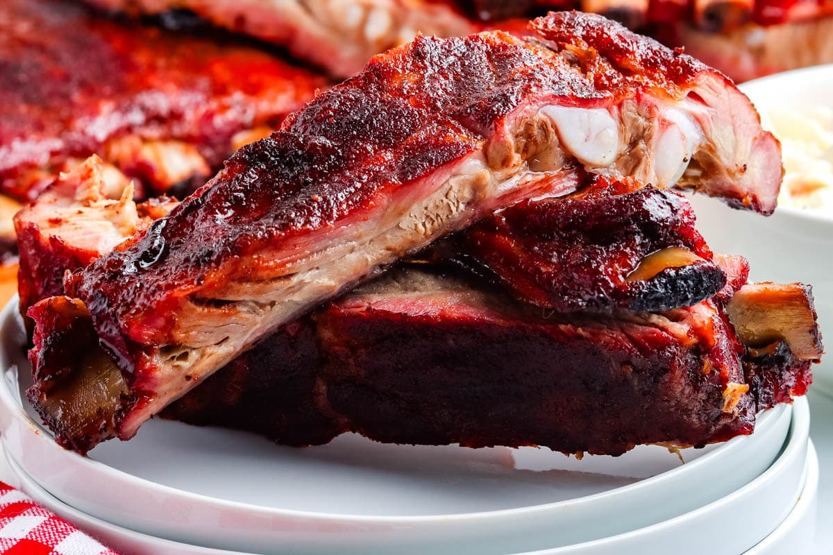 Two juicy, saucy barbecue pork ribs stacked on a white plate with more ribs in the background.