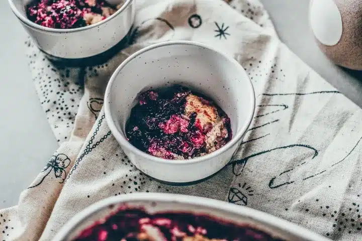 A bowl of berry crumble dessert on a patterned cloth, with two more bowls in the background.