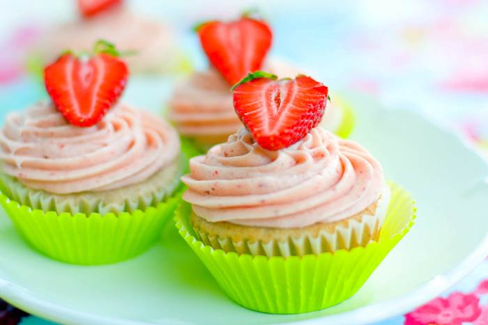 Cupcakes with pink frosting and halved strawberries on top, in green wrappers, on a white plate.
