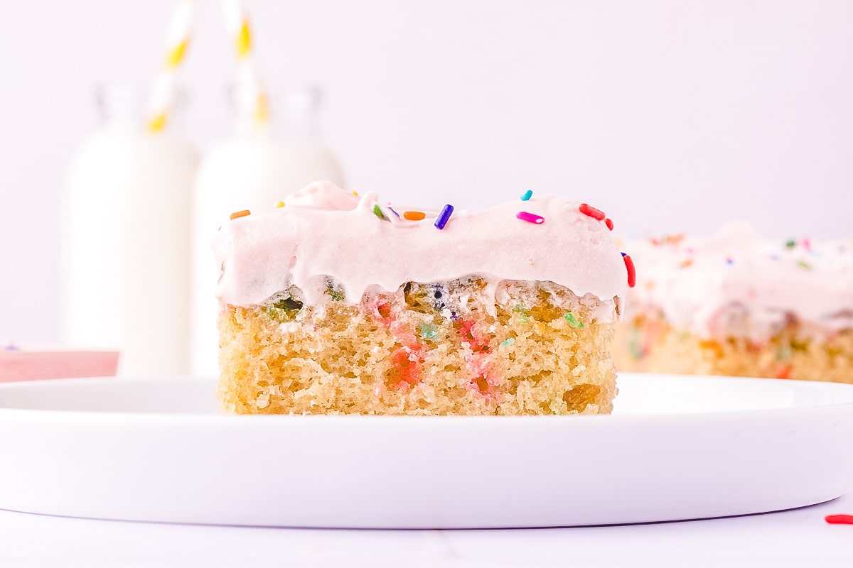 A slice of funfetti cake with pink frosting and sprinkles on a white plate, with milk bottles in the background.