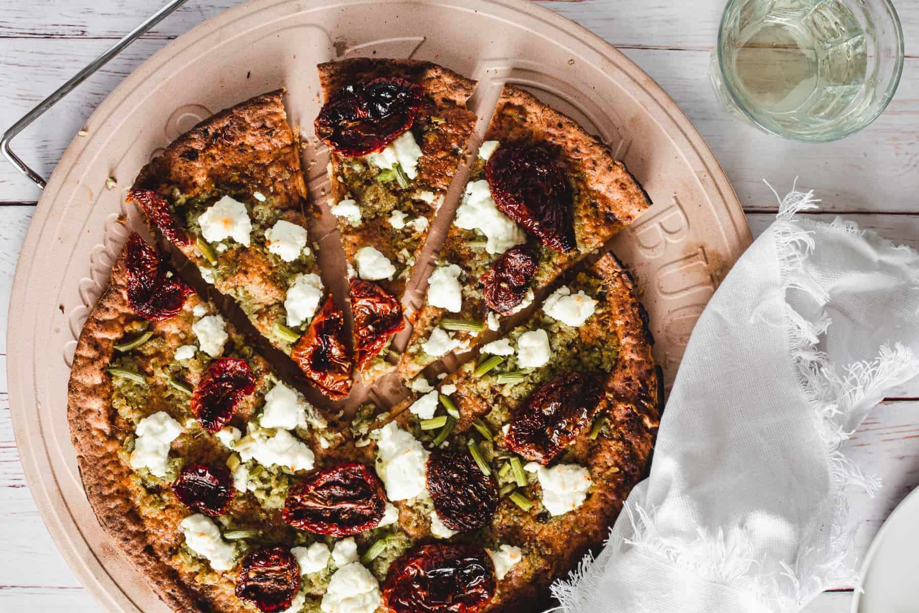 A sliced pizza with sun-dried tomatoes, goat cheese, and green toppings on a round tray next to a glass of water.