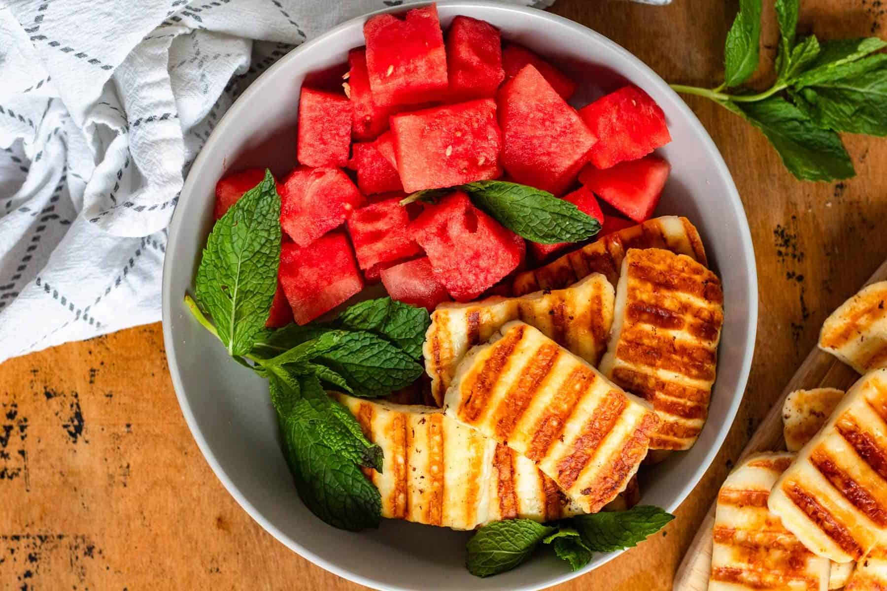 A bowl with grilled halloumi, cubed watermelon, and fresh mint leaves on a wooden surface.