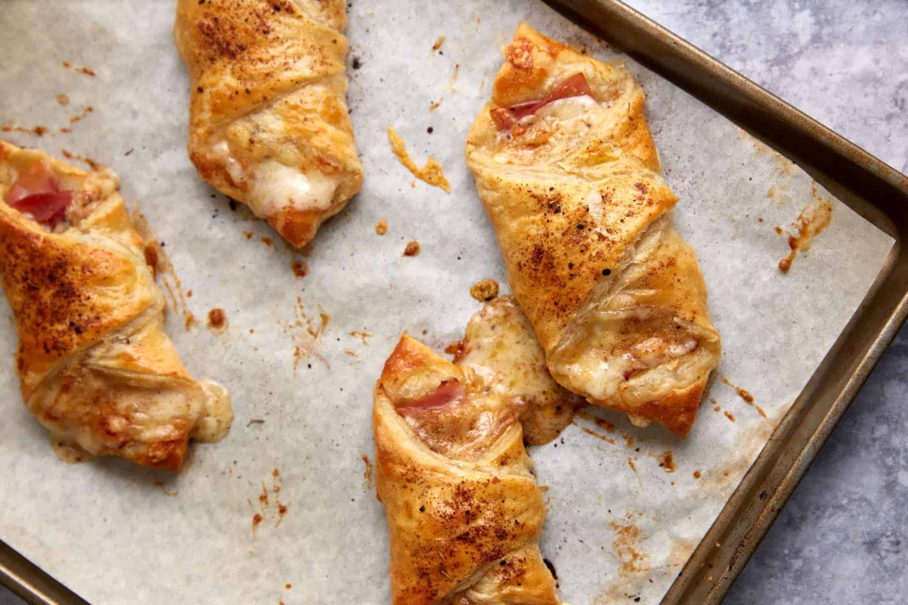 Four baked ham and cheese pastries on a parchment-lined baking sheet, with melted cheese oozing out.