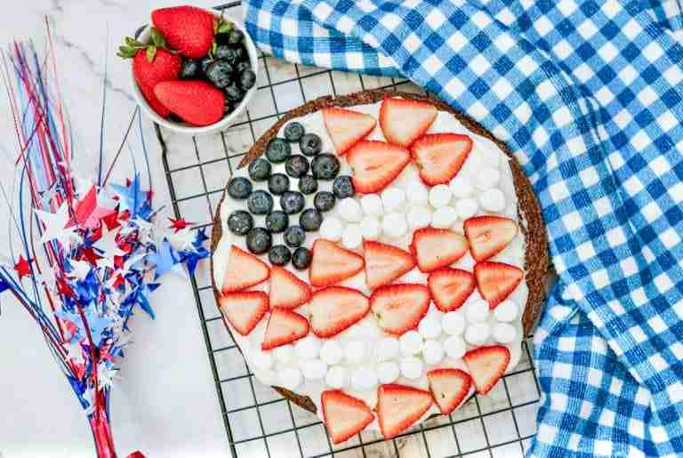 A dessert pizza decorated like an American flag with strawberries, blueberries, and marshmallows on a cooling rack.